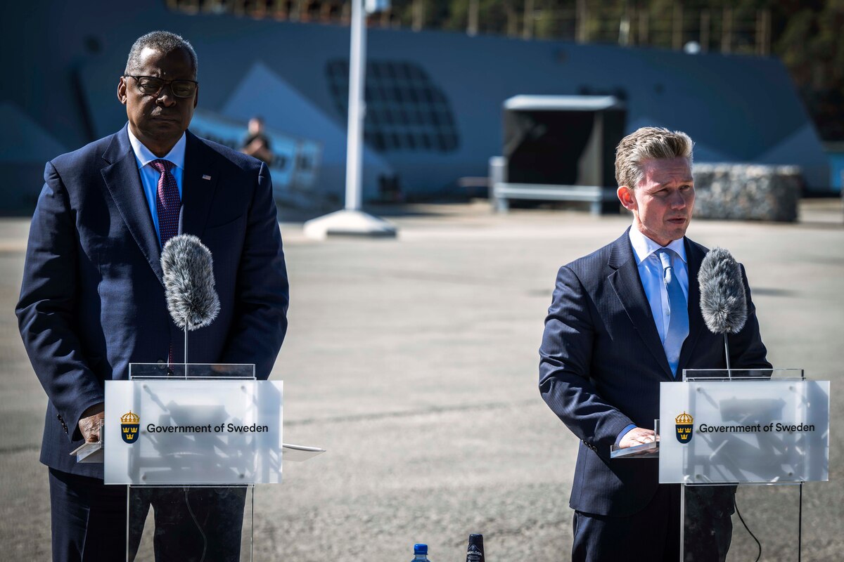 Two men stand behind lecterns. In the rear is a large naval vessel.