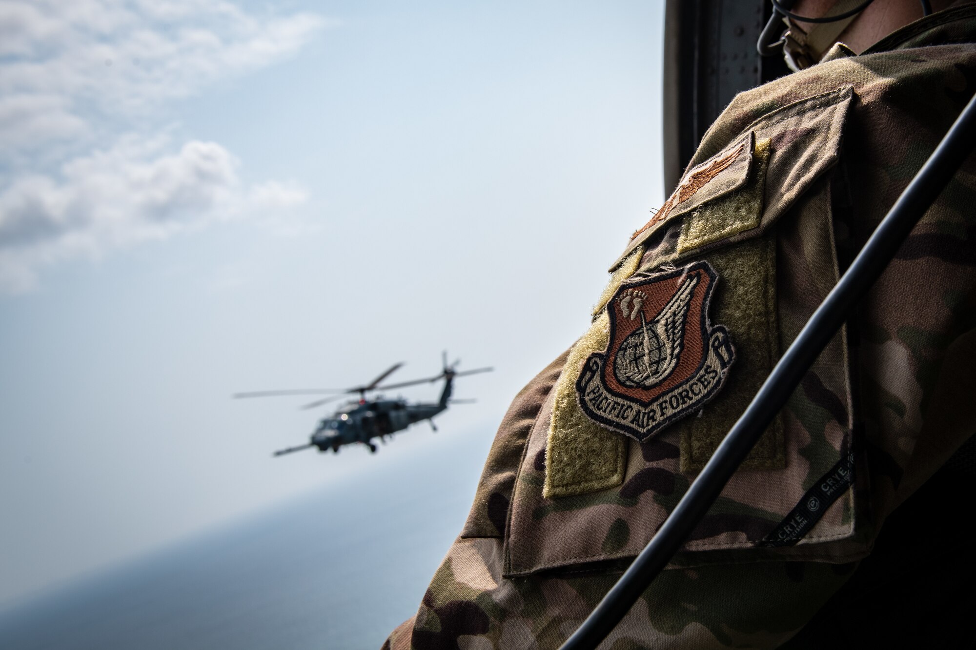 A U.S. Pacific Air Force patch on a uniform is in focus in the foreground while a helicopter flies over the water in the background slightly out of focus.