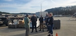 Group of people listening to man speak in scrap yard.