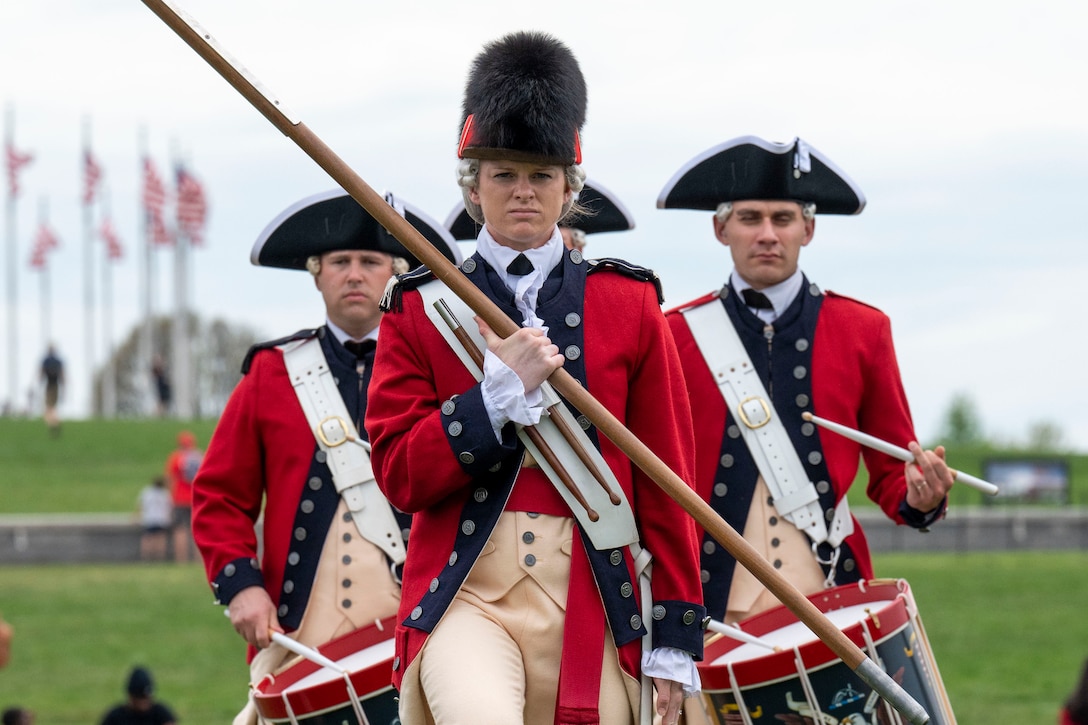 Three soldiers dressed in colonial uniforms march in formation.