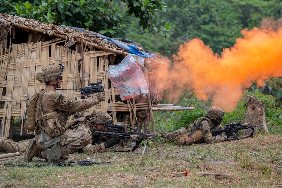 Soldiers with weapons kneel and lie on the ground as orange smoke floats through the air.