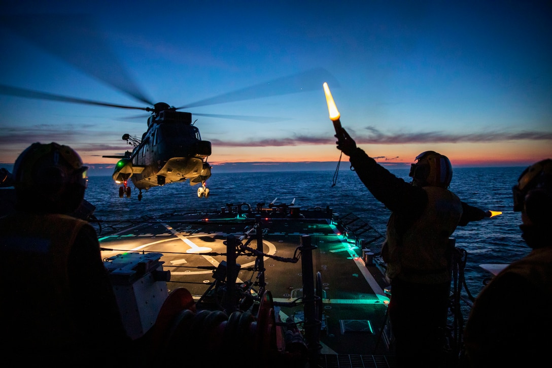 A sailor gives directions to a helicopter landing at night on a ship at sea.