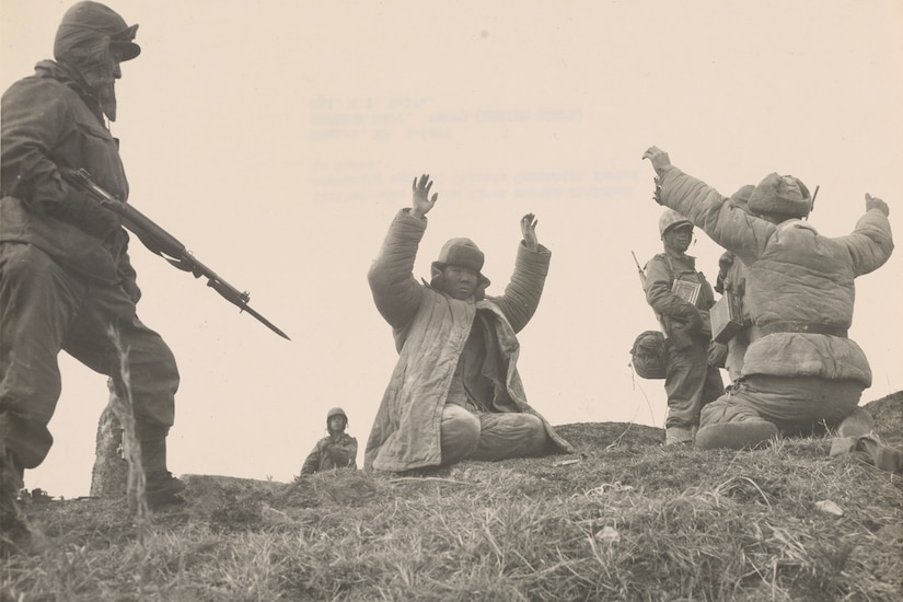 A man holding a rifle points it toward two kneeling men with their hands up in defeat.