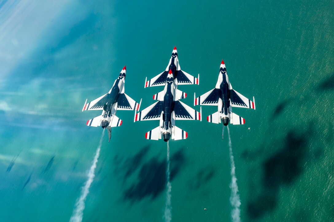 Four aircraft fly in formation over bluish green-water with cloud shadows.