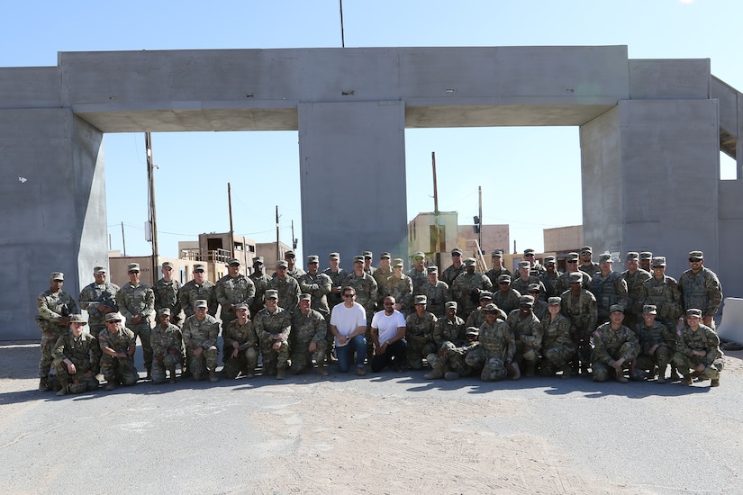 Two people and a large group of service members pose for a photo in front of a large concrete structure.