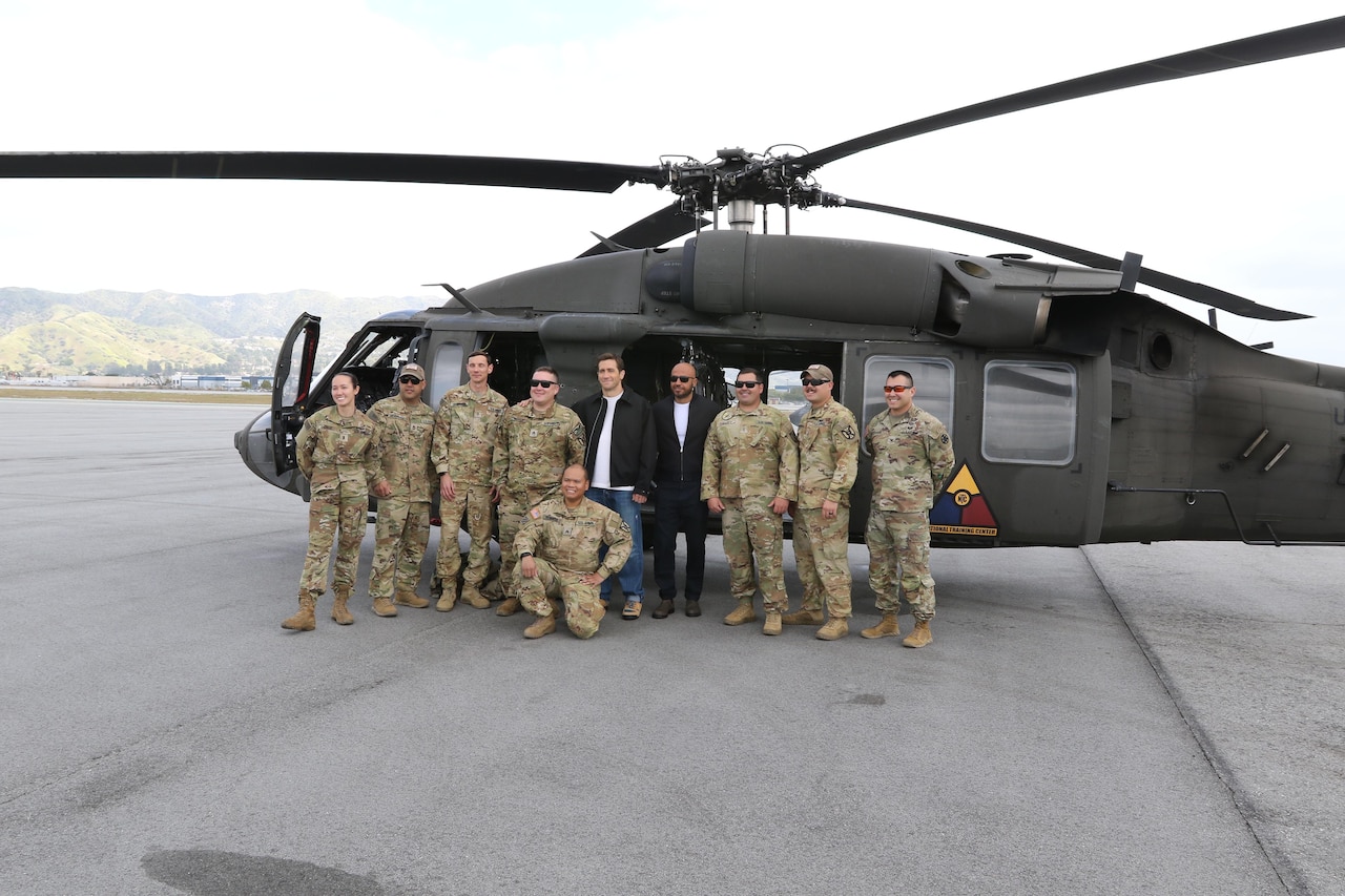 Two people stand with a group of uniformed service members in front of a helicopter.