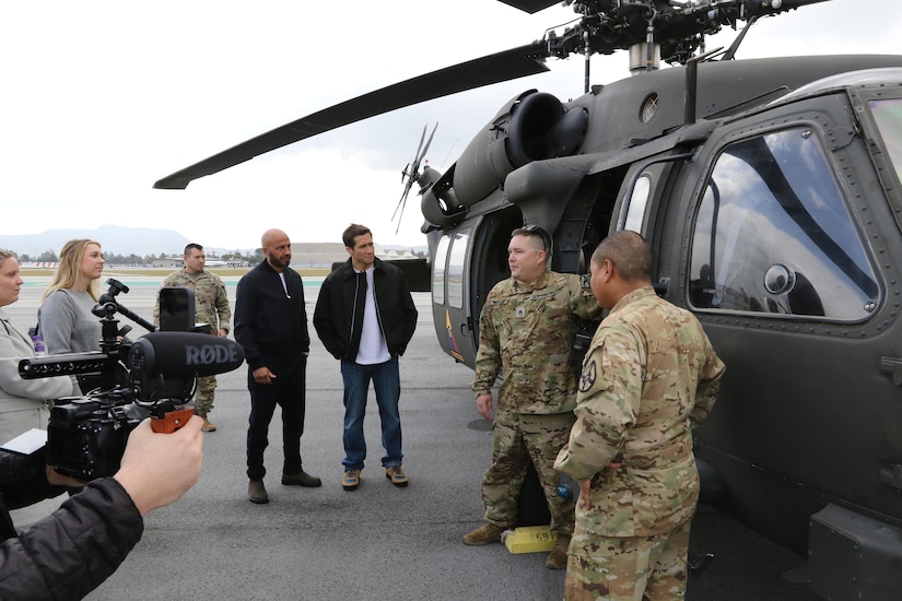 Two uniformed service members talk to a group of people  next to a helicopter.