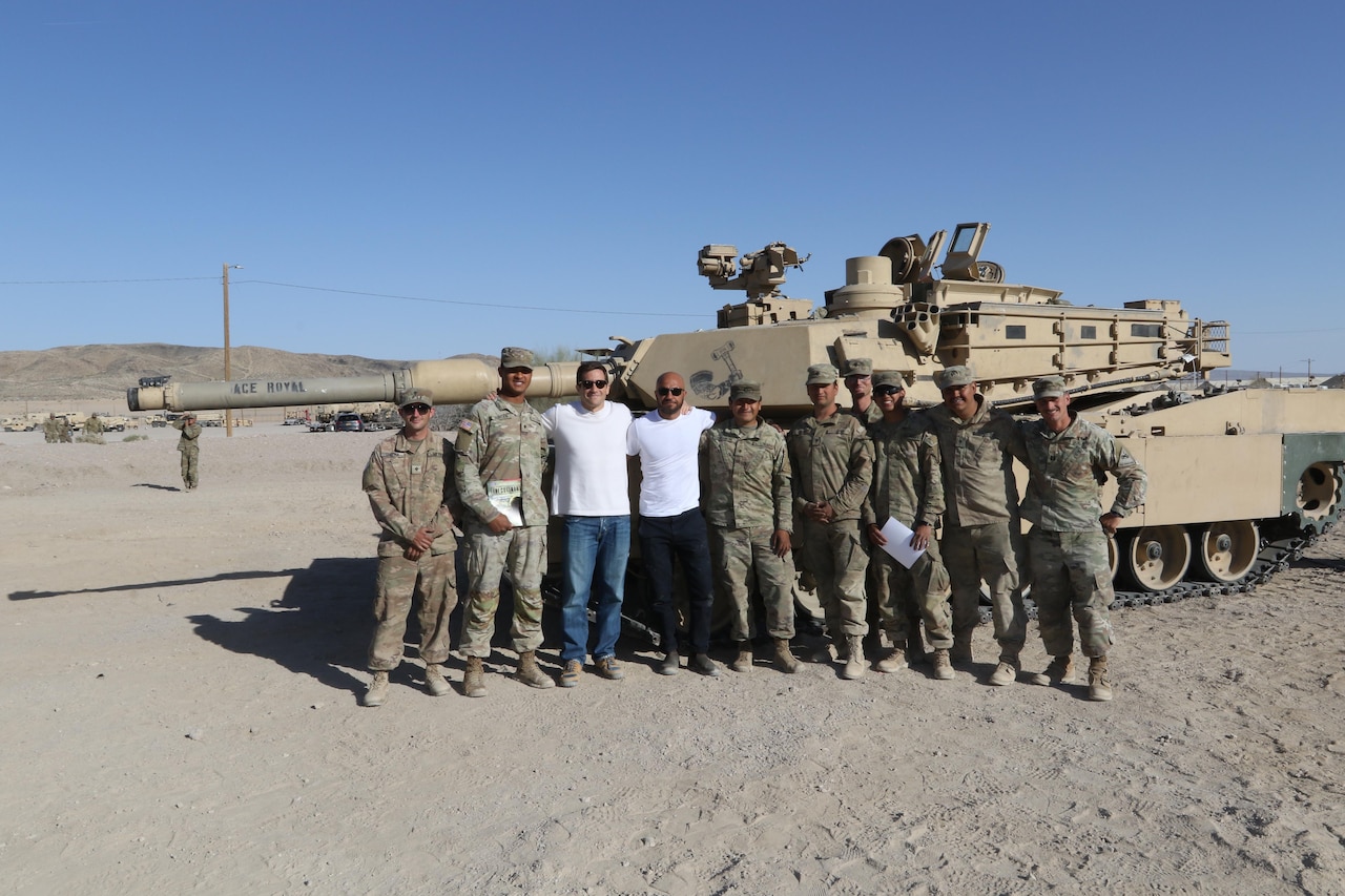 Two people and a group of uniformed service members pose in front of a tank in the sand.