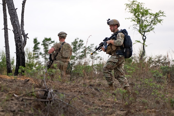 Two Airmen are holding weapons on a hill, one in the foreground walking towards the other who is kneeling while looking into the distance.