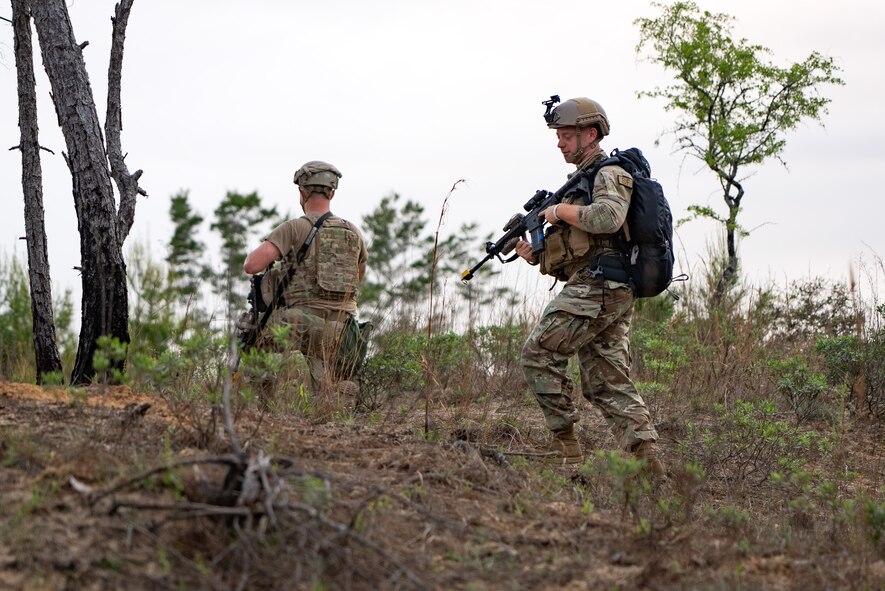 Two Airmen are holding weapons on a hill, one in the foreground walking towards the other who is kneeling while looking into the distance.
