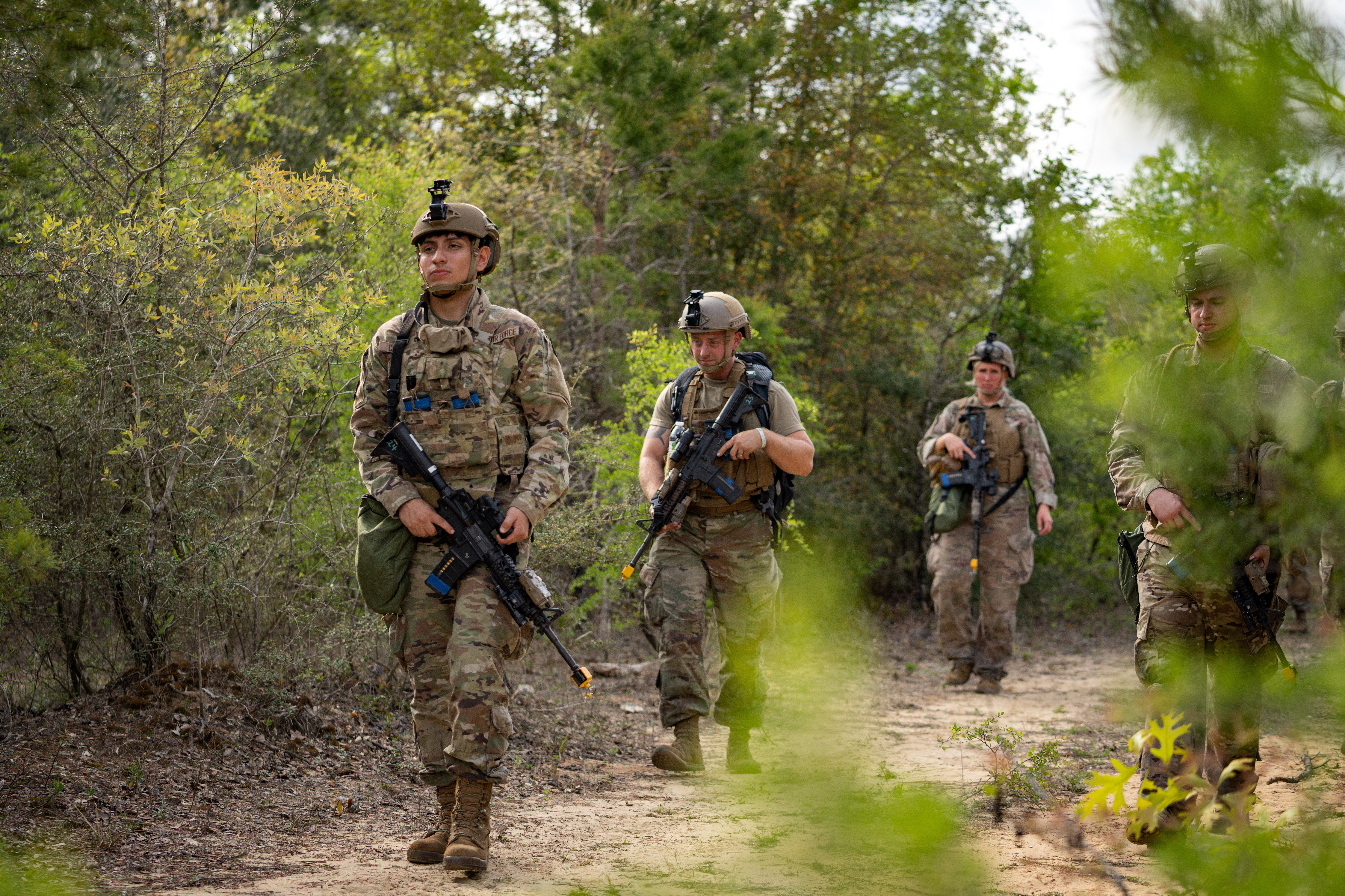 Four Airmen carrying weapons in tactical gear walk on a dirt path in the forest while spaced out in a defense formation