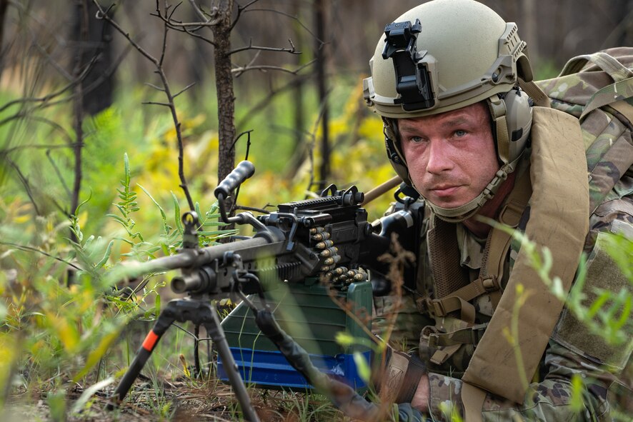 Airman with helmet and tactical gear is laying between ferns in the woods and handling a large weapon