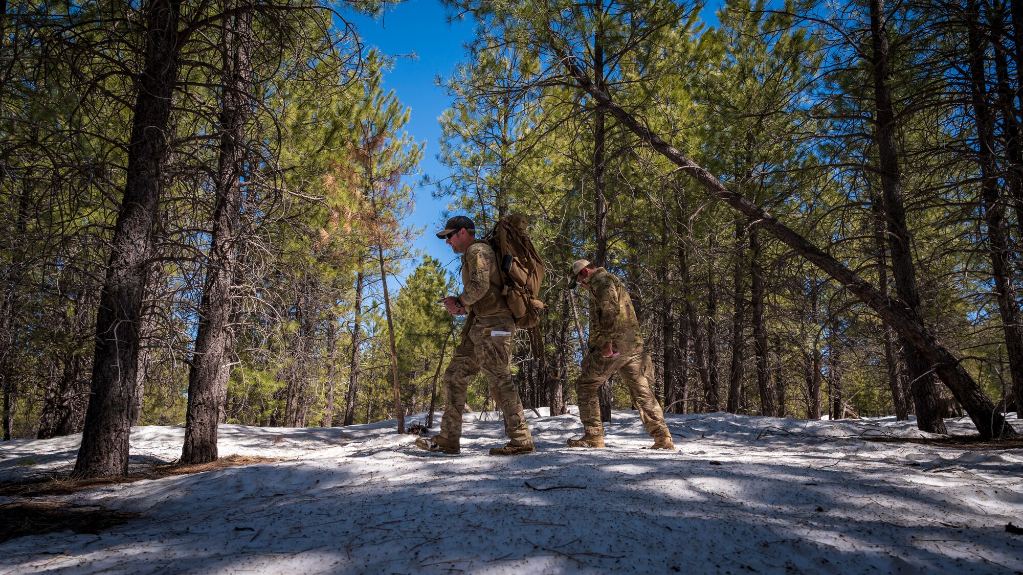 U.S. Air Force Tech. Sgt. Jason Evans (left) and Senior Airman Nestor Guevara-Penella (right), 56th Civil Engineer Squadron Explosive Ordnance Disposal flight technicians, hike through snow-covered pine forest at Camp Navajo, Arizona, April 11, 2023.