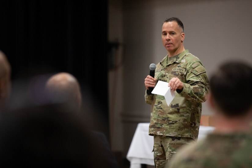 A Soldier standing and speaking in front of a sitting audience holding a wireless microphone and a piece of paper.
