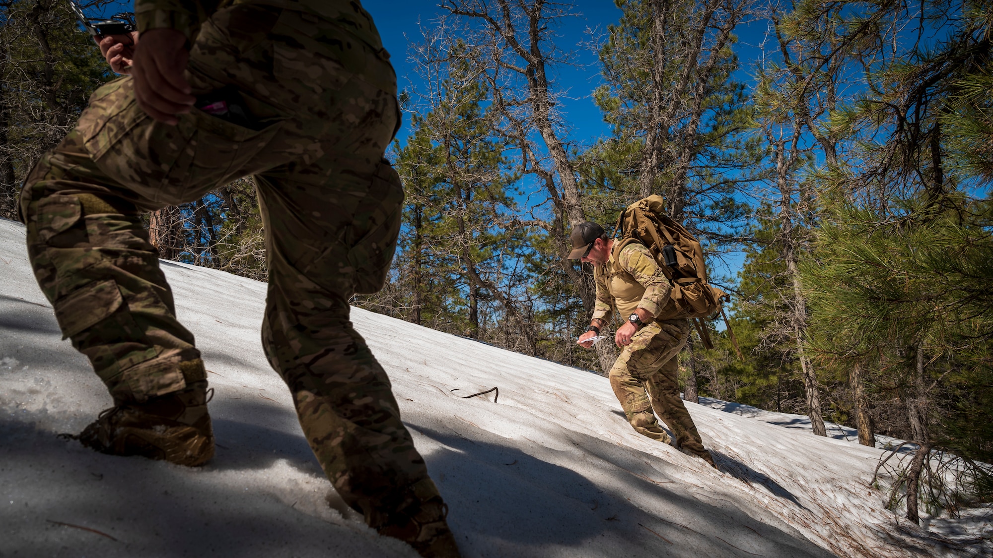 U.S. Air Force Tech. Sgt. Jason Evans (right) and Senior Airman Nestor Guevara-Penella (left), 56th Civil Engineer Squadron Explosive Ordnance Disposal flight technicians, hike up a snow-covered hill at Camp Navajo, Arizona, April 11, 2023.