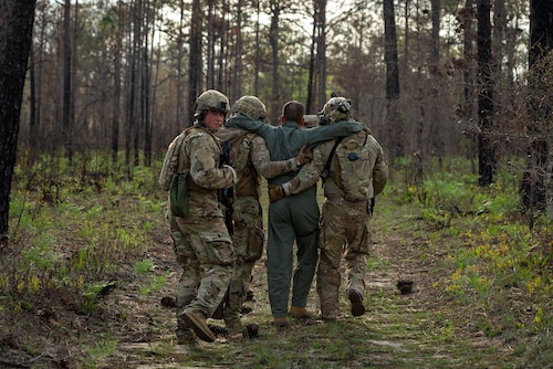 Two Airmen in OCP uniform carry another Airman in a flight suit who has his arms on their shoulders. An Airman just behind them is looking at the camera while holding a weapon.