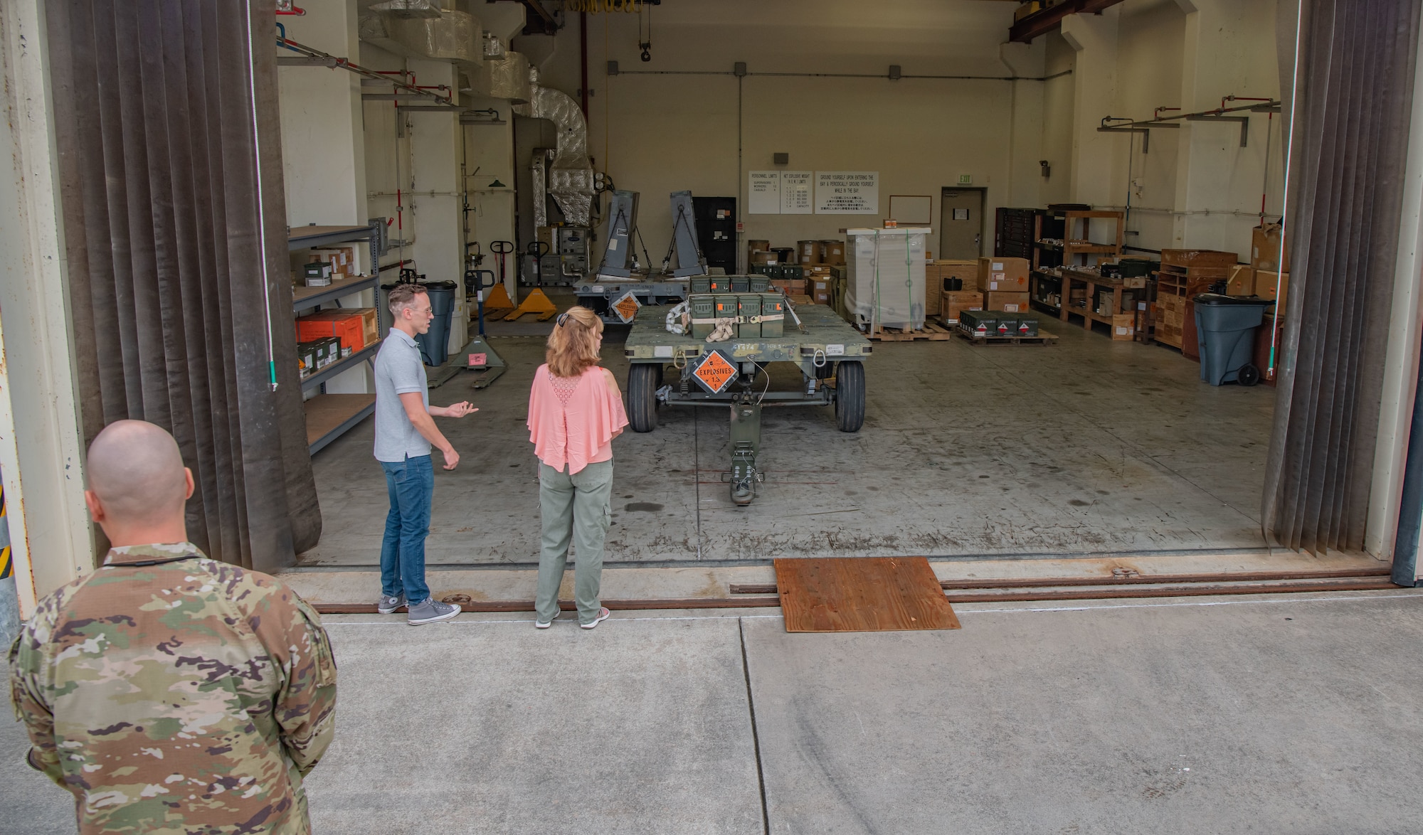 A man shows a woman a workstation containing a trailer, several boxes and munitions.