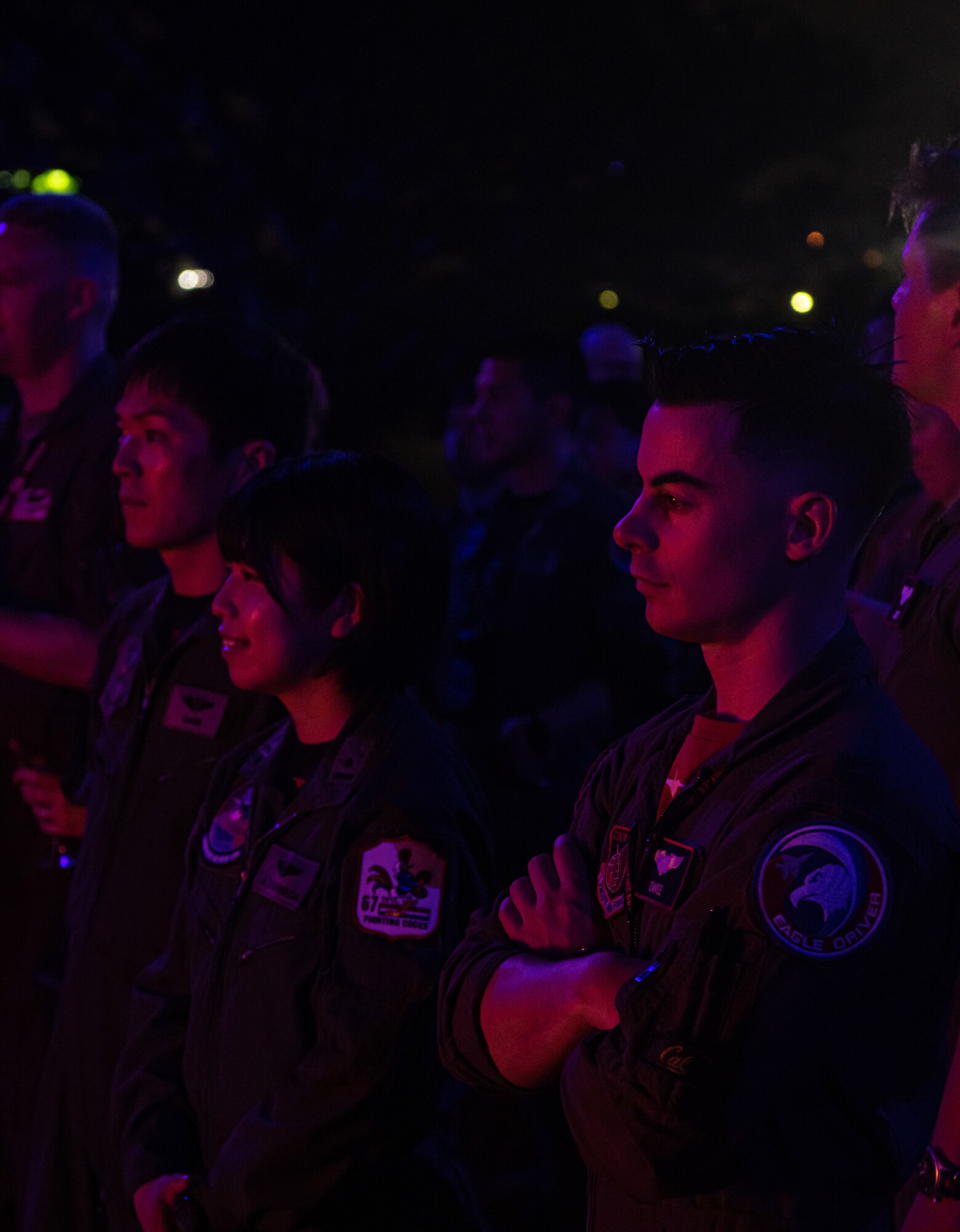 Three attendees of the piano burn watch a piano burn. The light is mixed with reds and blues from the streetlights and the fire.