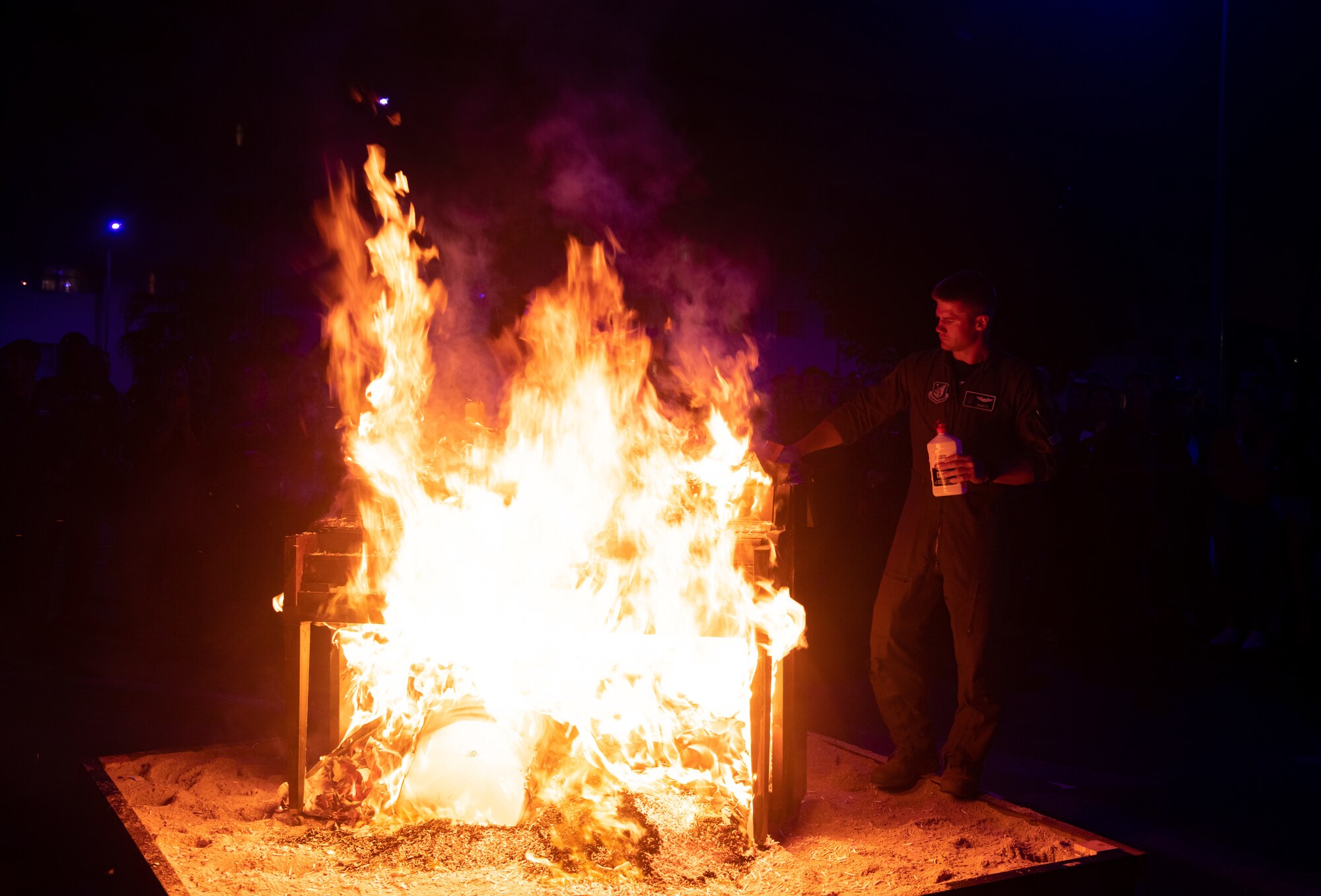 A pilot holding a bottle of lighter fluid adds kindling to a burning piano.