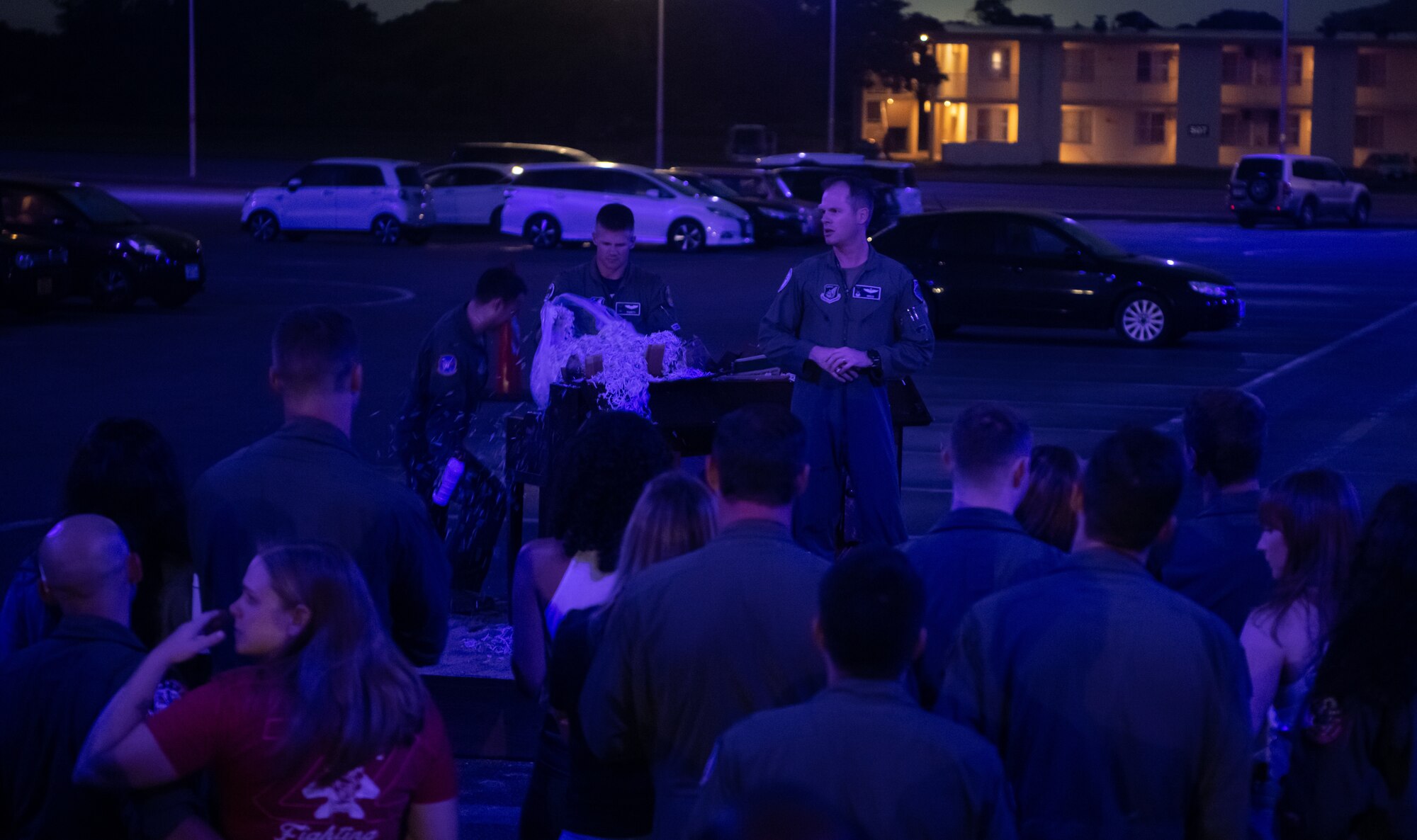A pilot talks to a crowd of people while two individuals in the background prepare to light a piano on fire.