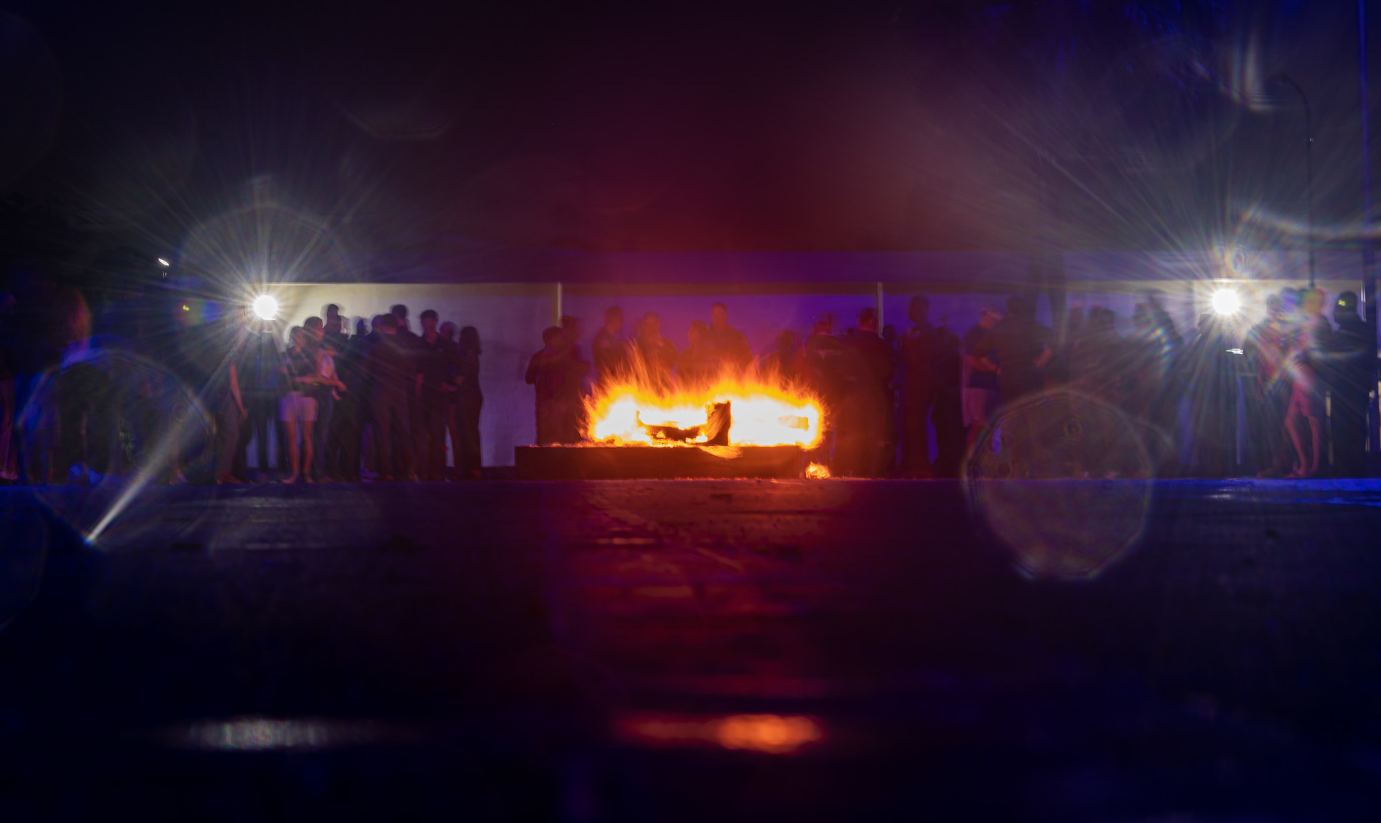People gather around a burning piano. Lights reflect back into the camera creating a starburst effect.
