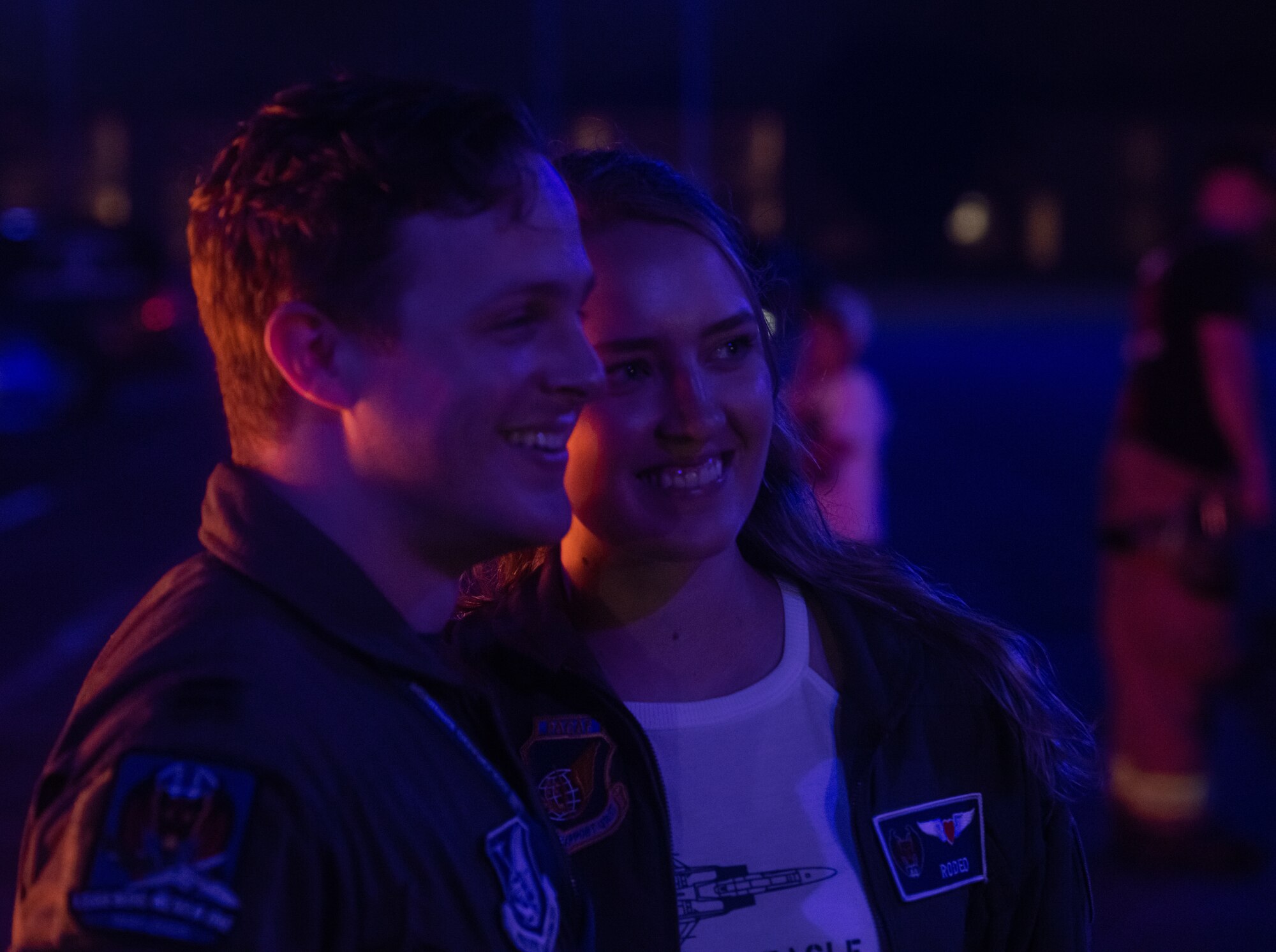 A man and a woman pose for a photo. Blue and red light is reflected on them from the streetlights and the burning piano.