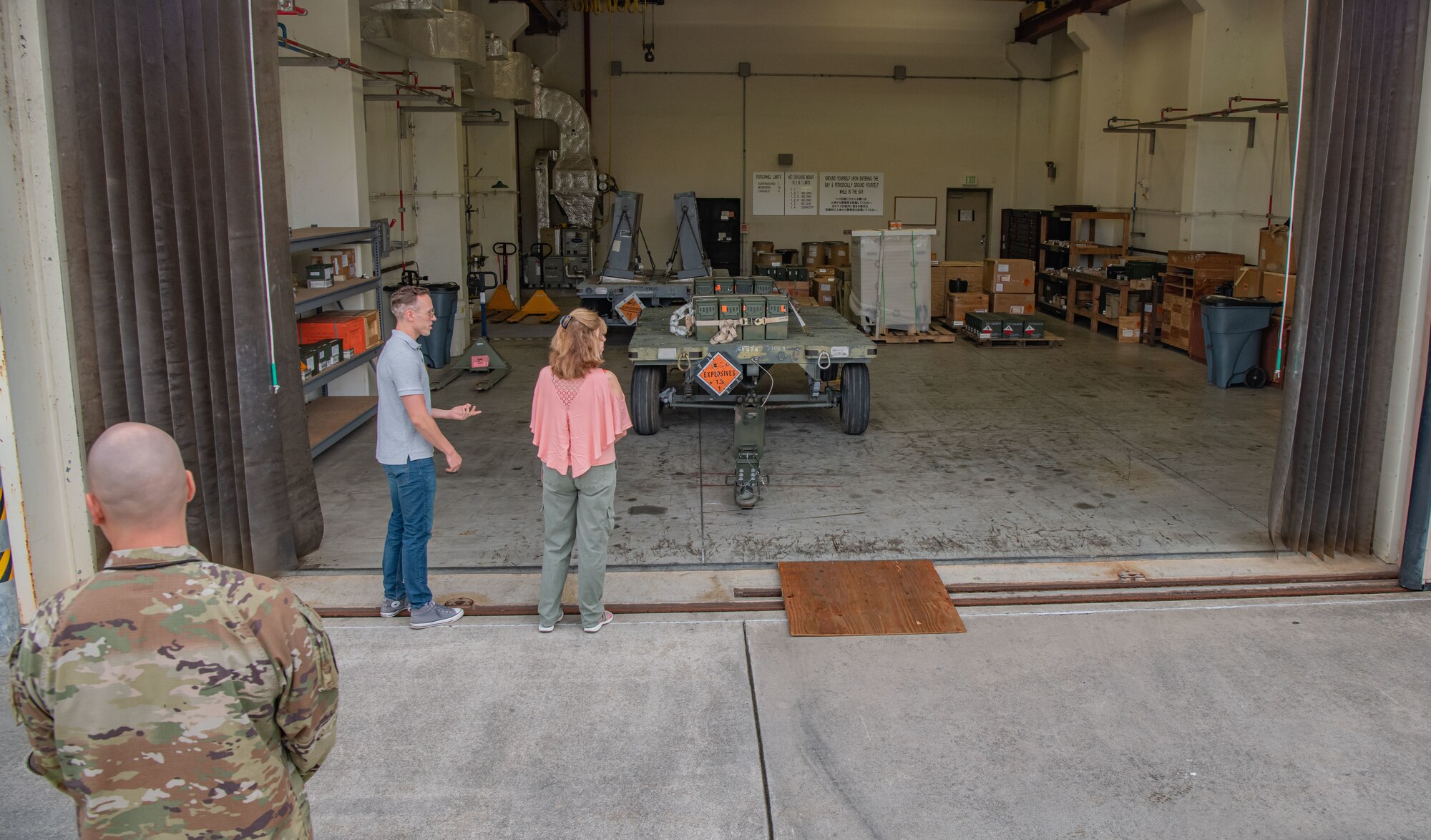 A man shows a woman a workstation containing a trailer, several boxes and munitions.