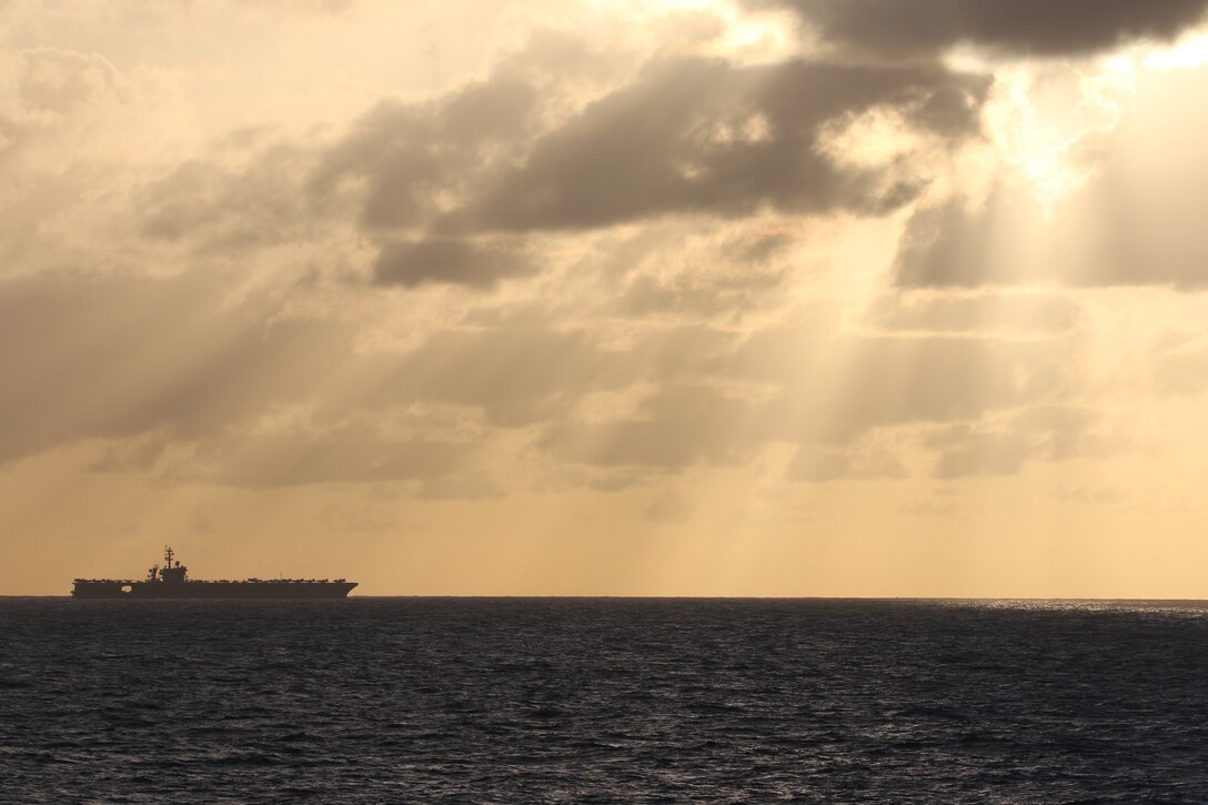 An aircraft carrier steams across a wide open ocean with filtered sunlight.