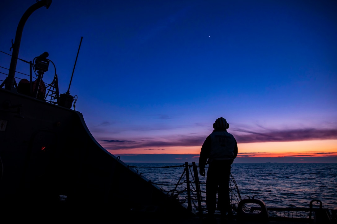 A sailor stands on the deck of a ship at twilight.