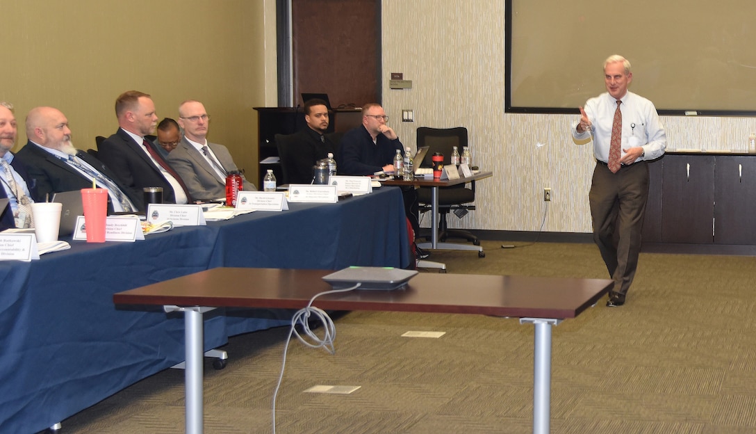 Man with white shirt standing in middle of conference room gesturing while speaking to seated conference participants.