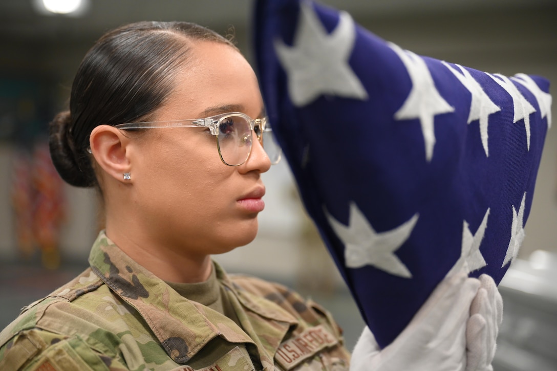 A service member wearing glasses and white gloves holds up a triangular-shaped folded American flag.