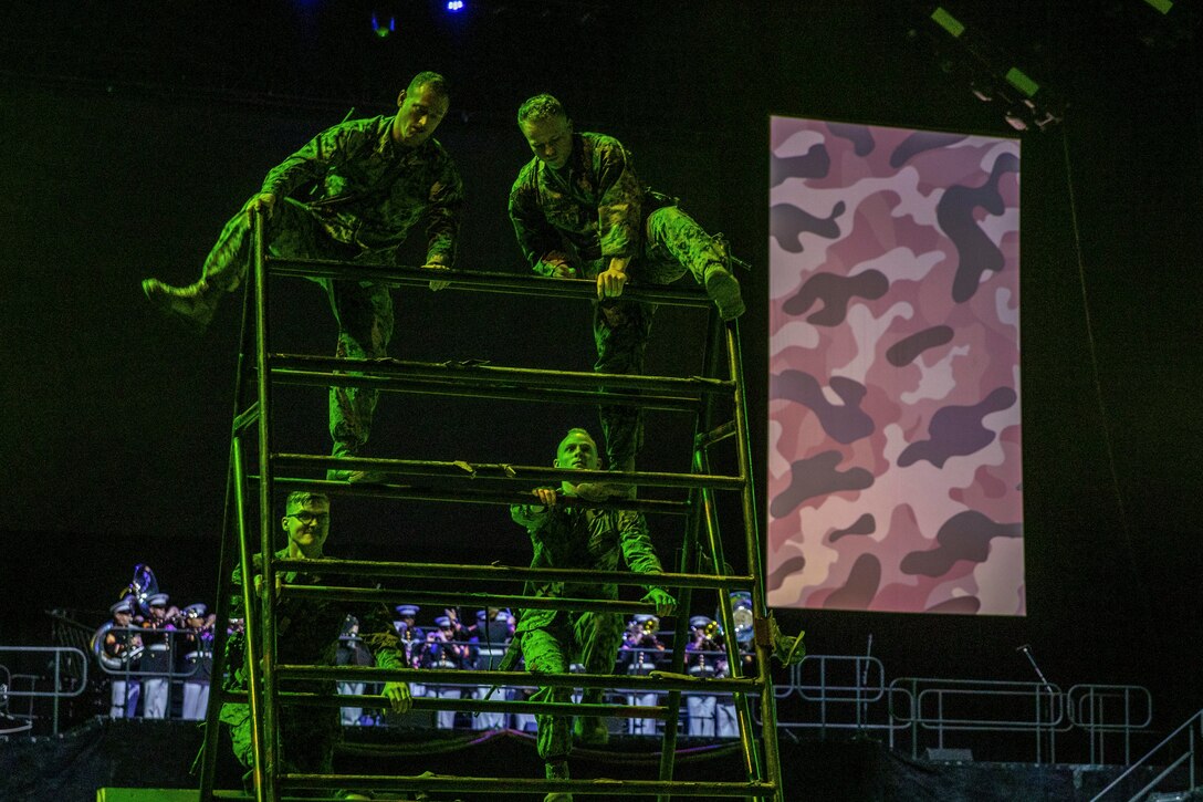 Four Marines climb an obstacle as a band performs in the background.