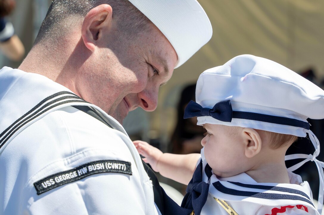 A sailor smiles while holding his infant daughter who is dressed in a sailor’s uniform.