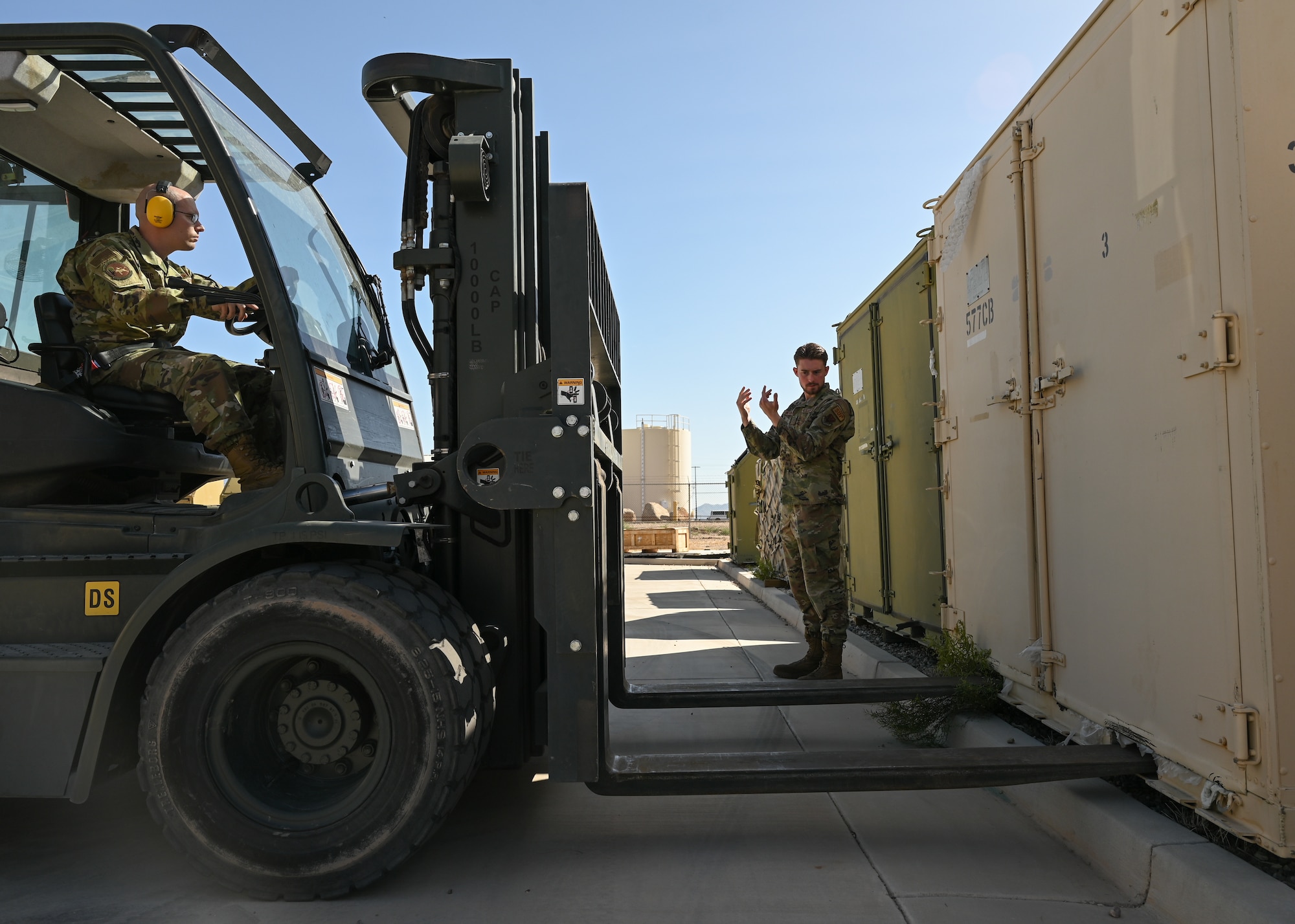 A man in a military uniform operates a forklift to pick up a large metal crate.