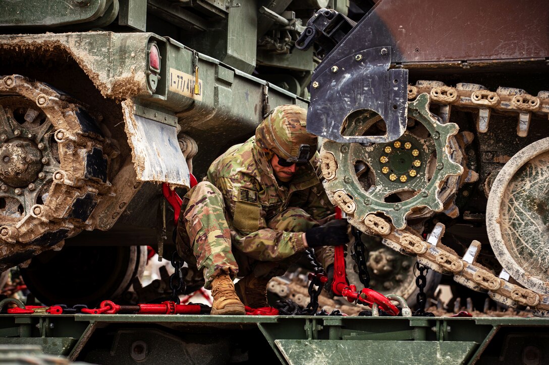 A soldier kneels while working on heavy equipment.