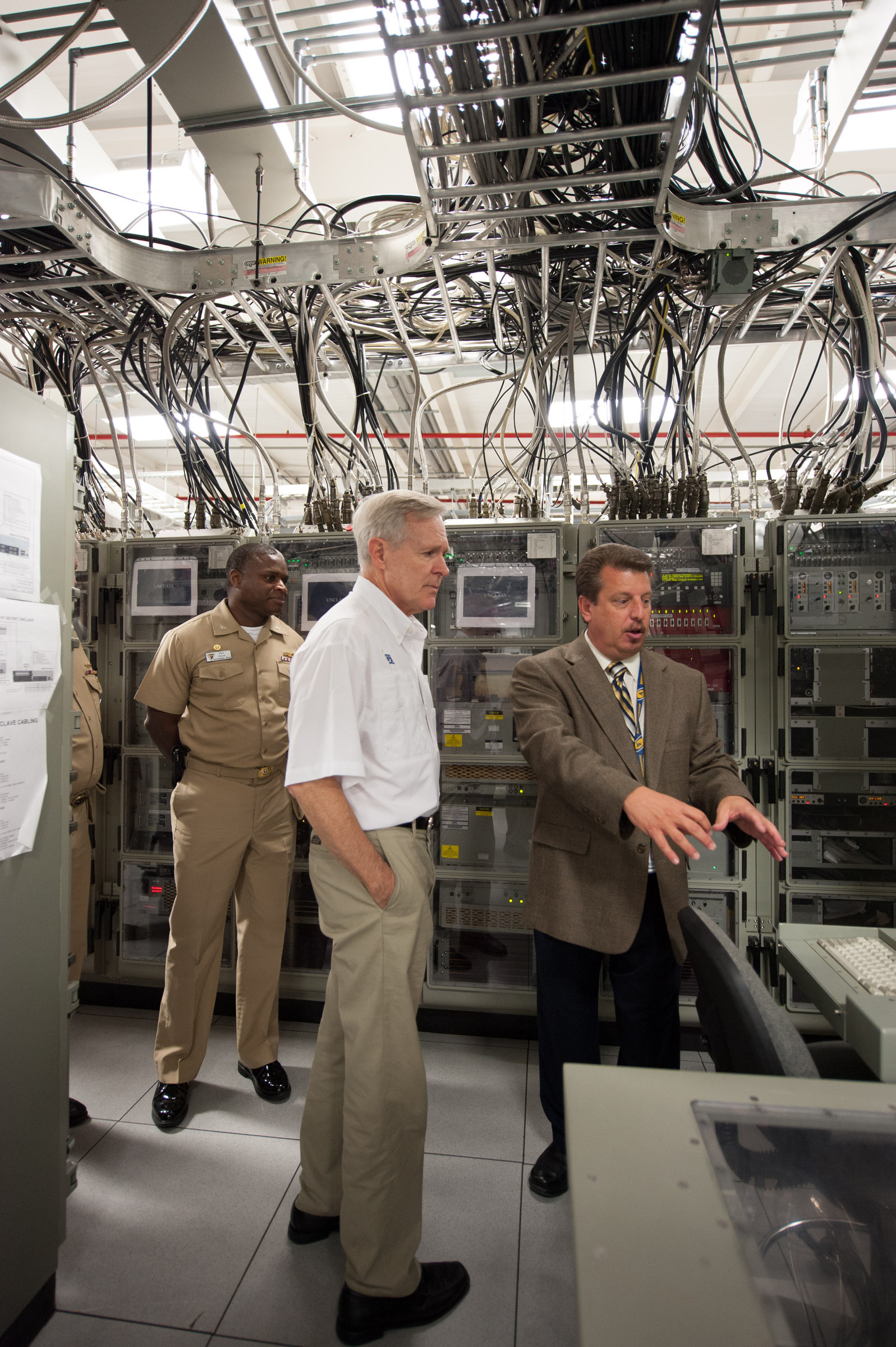 Secretary of the Navy looking at a radio room test are with a uniformed sailor and civilian.