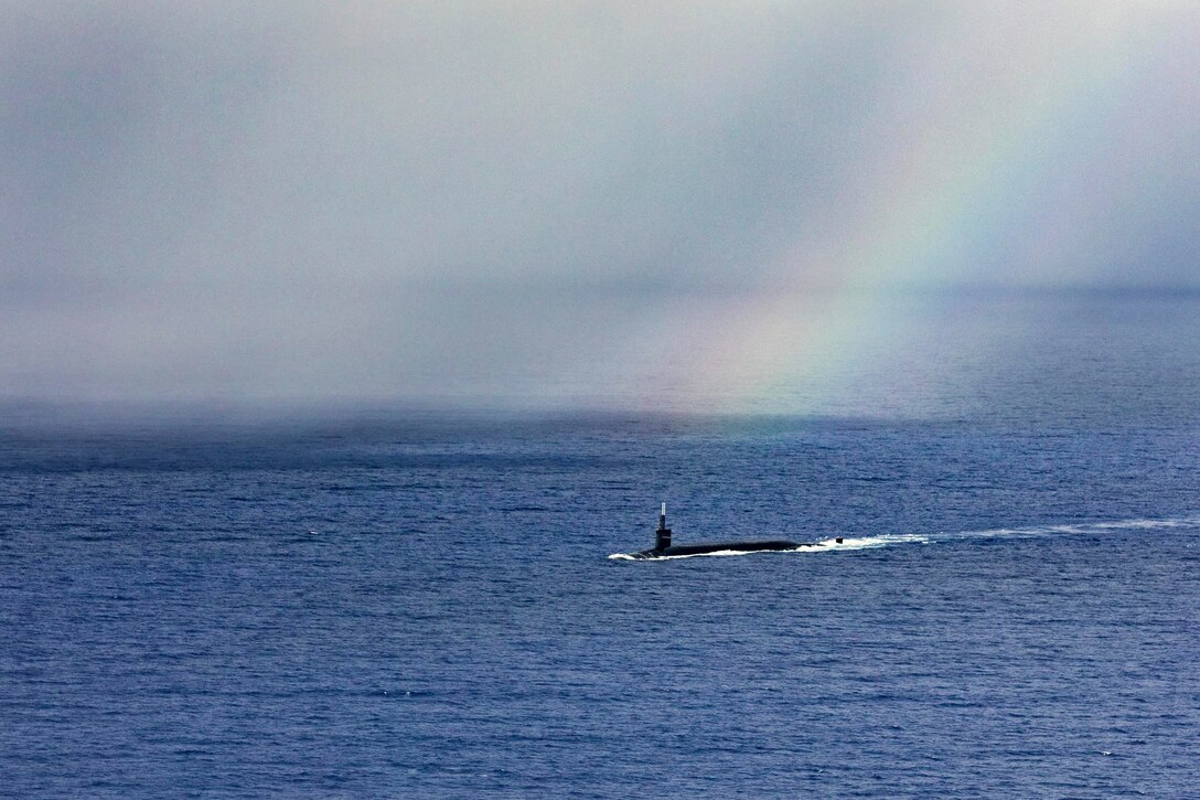 A submarine transits a body water under a rainbow.