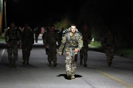 Soldiers walking in the middle of the street with ruck sacks in the early morning while dark out.