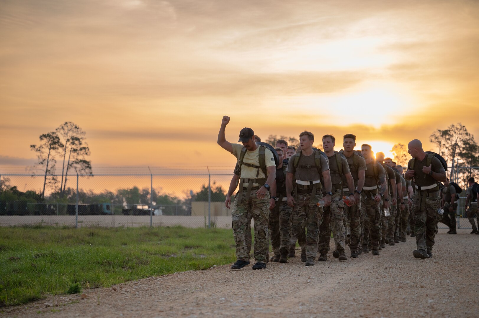 Instructor holds up arm to signify a stop to the group of students behind him as they ruck with weights on their back at sunrise