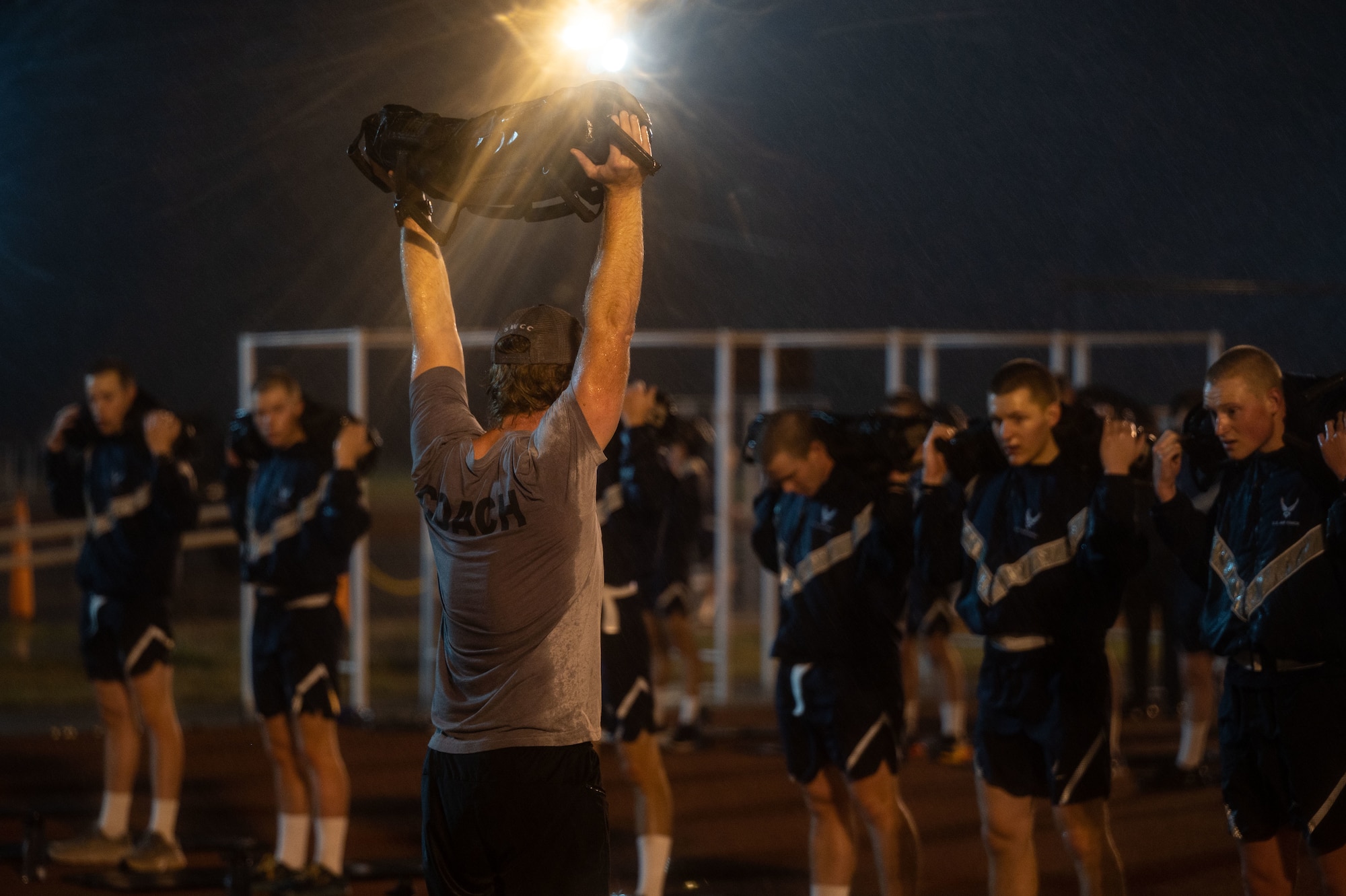 Coach demonstrates workout with weight over his head in front of group of students in the rain