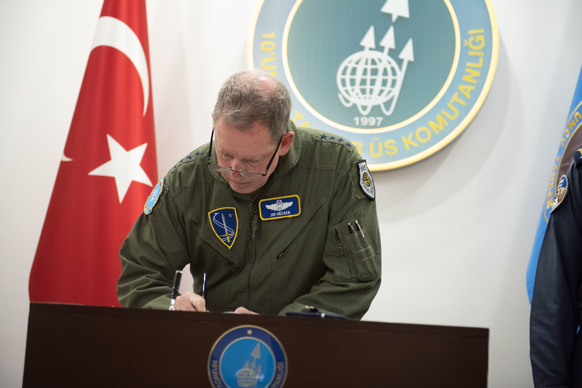 A man in a green flight suit signs a book placed on a podium.