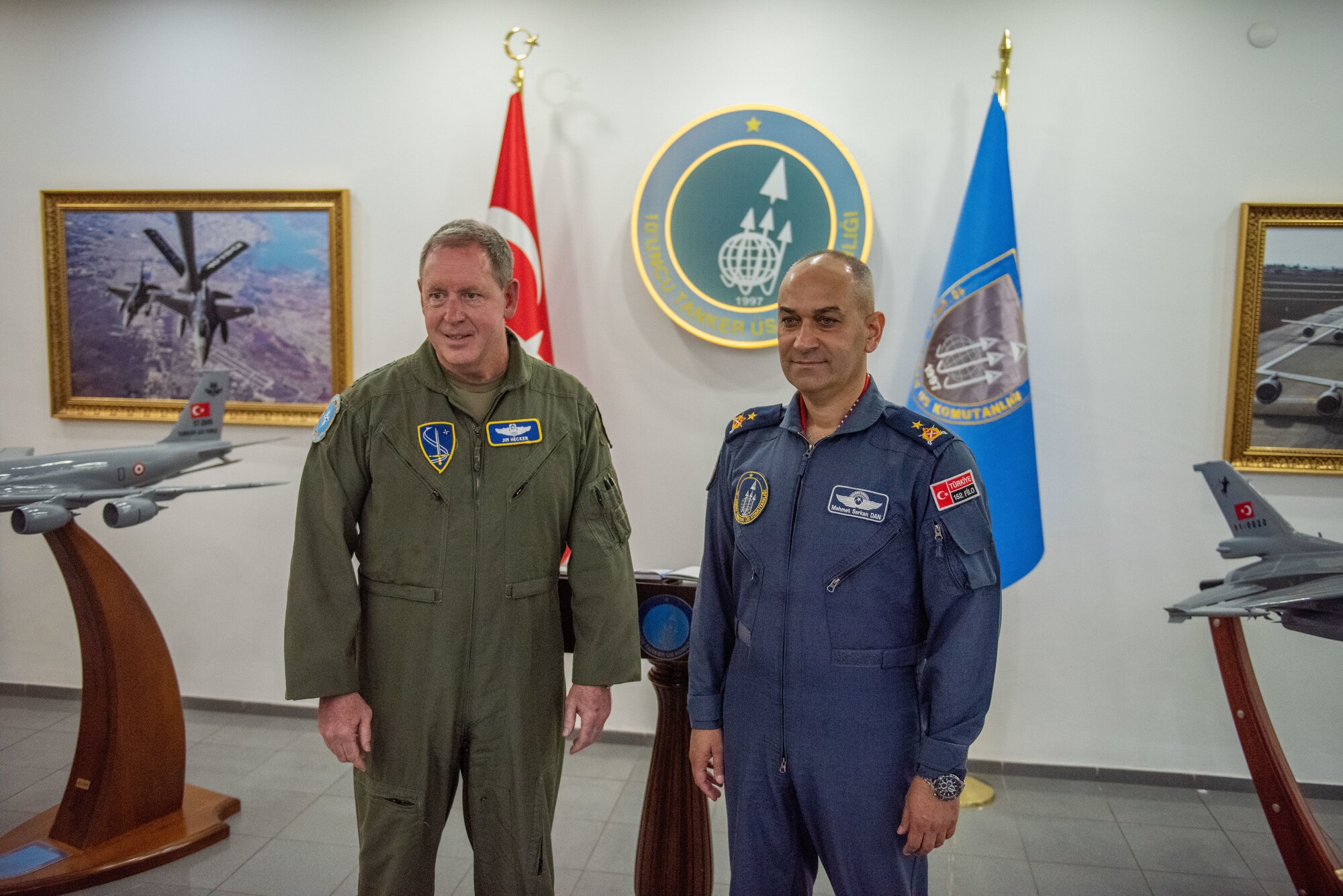 A man in a green flight suit stands next to a man in a blue flight suit. They're flanked by flags and model aircraft.