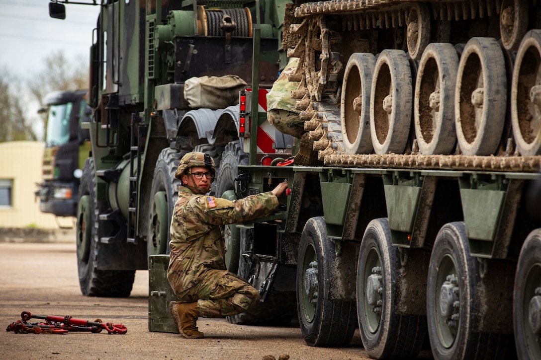 A soldier kneels beside heavy equipment that is loaded and ready for transport.