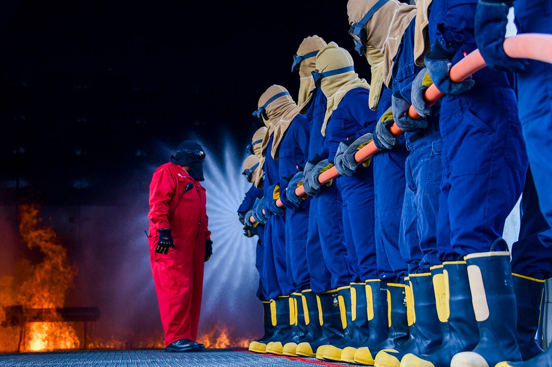 Sailors in protective gear stand in a line while holding a hose as they receive information from a trainer.