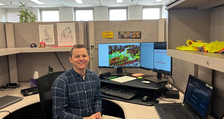 Man wearing blue and white plaid shirt sits at desk smiling.