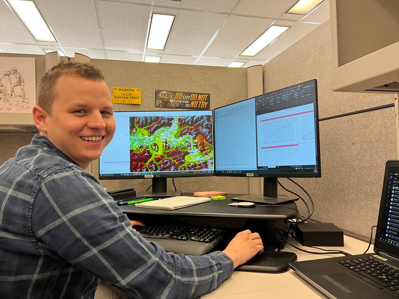 Man wearing blue and white plaid shirt sits at desk smiling.