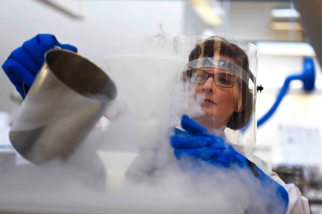 A scientist wearing personal protective equipment pours liquid from a canister as smoke rises.