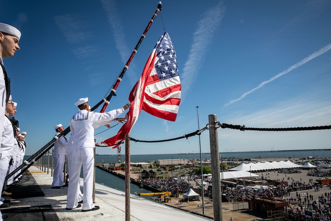 Sailors raise a flag about a ship as a crowd gathers on the ground nearby.