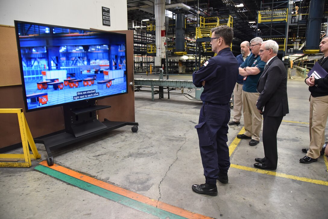 group of men standing in a warehouse watch a video of modernization efforts