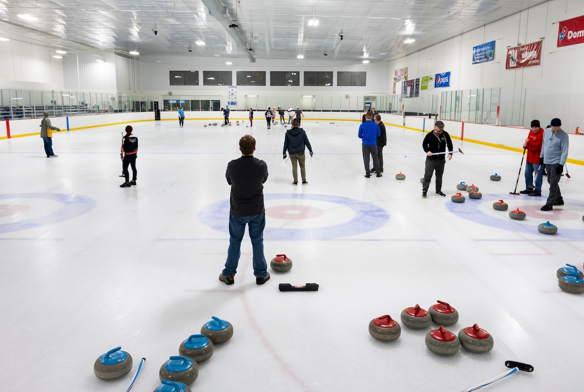 People stand scattered across a hockey rink. Curling stones are arranged in the foreground.